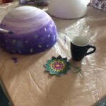 Foto: auf eine Tisch eine blau bemalte Styropor-Halbkugel und eine malende Hand mit Pinsel im Vordergrund, eine weiße Styropor-Halbkugel und Malzeug dahinter.