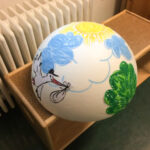 Foto: eine mit Sonne und Wolke bemalte und mit Baum und Storch sowie Wolken beklebte Styropor-Halbkugeln auf einer Bank vor einem Heizkörper.