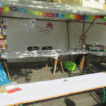 Foto: Menschenleerer überdachter Stand des FZM mit Prospekten darauf und Logo-Banner an Rückwand. Im Vordergrund Tisch zum bemalen.