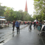 Foto: viele Menschen laufen auf regennasser Strasse zwischen überdachten Ständen. Kirchturmspitze im Hintergrund.