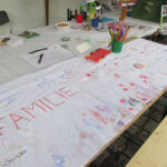 Foto: Tisch diagonal im Bild v.l.u.n.r.o. mit bunt bemalten zusammengeklebten Papierblättern und Malstiften darauf. Tisch mit Prospekten im Hintergrund.