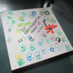 Foto: weiße Holzplatte mit unterschiedlich farbigen Handabdrücken, dem FZM-Logo und dem Schriftzug "Familinezentrum Moabit-Ost ist bund!"