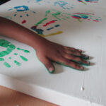 Foto: weiße Holzplatte mit unterschiedlich farbigen Handabdrücken und einer Hand die gerade einen grünen Abdruck macht.