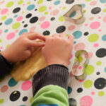 Foto: Kinderhände beimausrollen von Plätzenteig auf einer bunt bepunkteten Tischdecke