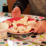 Foto: Kinderhände beim verzieren von Plätzen mit Zuckerguß und bunten Streuseln auf einem Teller auf einer bunt bepunkteten Tischdecke