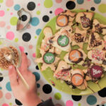 Foto: Kinderhände beim verzieren von Plätzen mit Zuckerguß und bunten Streuseln auf einem Teller auf einer bunt bepunkteten Tischdecke