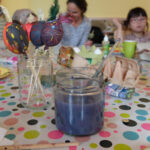 Foto: im Vordergrund Malfarben und Glas mit Pinselwasser und Pinsel sowie bunt bemalte Eier am Holzstil in eine Glas. Im Hintergrund Kinder und eltern beim bemalen von Eiern.