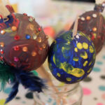 Foto: 3 ausgeblasene Eier, bunt bemalt und teilweise mit bunten Glassteinchen und Federn beklebt, stehen jeweils am Holzstil in einem Glas.