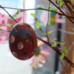 Foto: ein bunt bemaltes ausgeblasenes Ei hängt an Zweigen mit kleinen grünen Blättern vor einer Fensterscheibe im Hintergrund.
