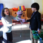 Foto: Frau knieend auf dem Fußboden mit einer Handpuppe auf dem Arm, führt die Hand der Puppe zum Handgruß mit einem Jungen.