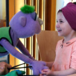 Foto: Handpuppe und Mädchen auf Stuhl sitzende halten gemeinsam beide Hände.