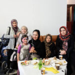 Fünf Jahre Familienzentrum Moabit-Ost - Glückliche Gäste | Foto: © FZM Foto: Gruppe von Frauen mit Kopftüchern und Kind mittendrin sitzen hinter einem Tisch mit Gebäck auf einer Couch und lächeln.