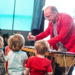 Foto: älterer Herr mit rotem Jacket steht hinter einem Vibraphon und reicht den davor stehenden Kinder einen Rasierpinsel.