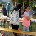 Foto: Frau mit Kopftuch und Kinder belegen Pizzateig. Im Hintergrund Mann und am Steinofen tätig.