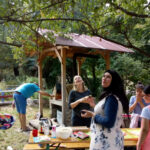 Foto: Frau mit Kopftuch und ohne schauen in die Luft. Daneben belegen 2 Mädchen Pizzateig. Im Hintergrund Mann am Steinofen tätig.