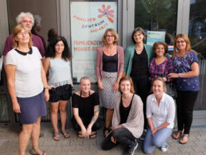 Foto: 9 Frauen und ein Mann stehen oder knien lose im Halbkreis vor der Eingangstür mit Logo des FZM.