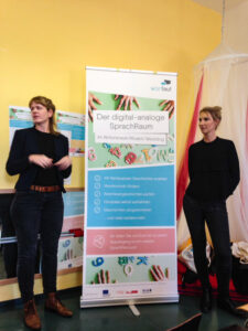Foto: zwei junge Frauen stehen, nach links blickend, jeweils rechts und links vor neben einer bunten beschrifteten Flipchart mit der Überschrift "Der digital-analoge SprachRaum"