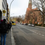 Foto: Blick von einer Bushaltestelle mit Haltestellen Schild und wartenden Reisenden. Buss rollt in der Ferne an.
