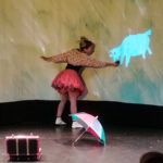 Foto: Blick auf die Bühne - Tänzerin hält einer Schafabbildung auf der Leinwand etwas zu fressen hin