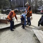 Foto: 2 Erwachsene in Warnwesten mit Müllbeutel und ein Kind beim Müll sammeln auf einem Spielplatz