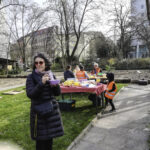 Foto: beim Pausieren - Erwachsene und Kinder am Esstisch im Hintergrund, eine Frau mit Essschüssel in der Hand im Vordergrund