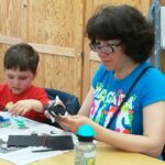 Foto: Frau mit blauen T-Shirt und Kind mit rotem Pullover sitzen an einem Tisch und basteln