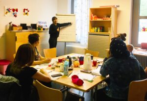 Foto: Raum mit Tisch in der Mitte an dem fünf Frauen sitzen während eine sechste Frau an einem Whiteboard etwas zeigt