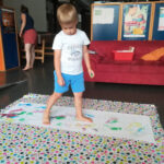 Foto: Eine bunte Decke liegt auf dem Boden, darauf ein Blatt mit aufgemalten Fußabdrücken, auf dem ein Junge steht.