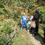 Foto: Ein Garten, links Sonnenblumen und Tomaten, rechts eine Frau mit Kopftuch und drei Kinder