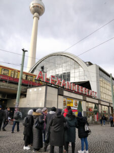 Foto: Brücke mit Zug, davor Schriftzug Alexanderplatz, unten steht eine Gruppe Frauen