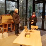 Foto: Raum mit großen Glasfenstern, im Hintergrund ein Regal, vorne ein Holztisch, dahinter stehen ein Mann und eine Frau mit Maske