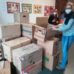 Foto: Viele gestapelte braune Umzugskartons, lins daneben zwei Fraun mit Maske