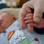 Foto: Neugeborenes Baby liegt und hält die Hand einer erwachsenen Person.