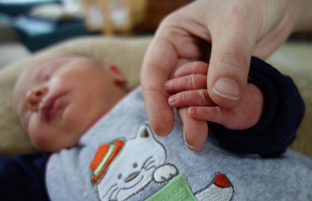 Foto: Neugeborenes Baby liegt und hält die Hand einer erwachsenen Person.
