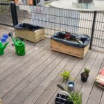 Foto: Eine Holzveranda, hinten zwei Blumenkästen, vorne Gartenhilfsmittel und frische Pflänzchen
