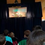 Foto: in der oberen Hälfte ein Puppentheater, unten sitzen fünf Kinder, die zuschauen