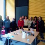 Foto: Im Vordergrund ein Tisch mit Kaffee und Gläsern, dahinter eine Gruppe von Menschen, die in die Kamera lächelt